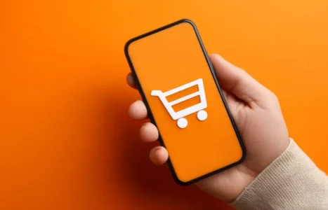 A hand holding a smartphone showing a white shopping trolley icon on an orange screen, illustrating AI assistant shopping recommendations for UK online shops.