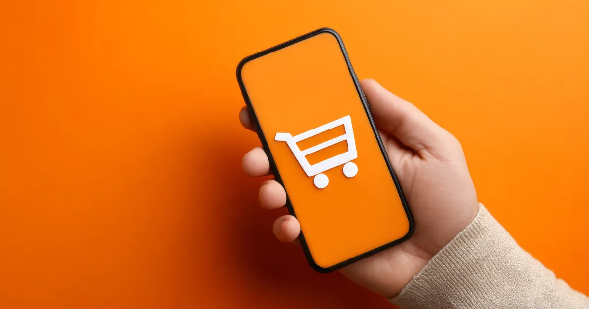 A hand holding a smartphone showing a white shopping trolley icon on an orange screen, illustrating AI assistant shopping recommendations for UK online shops.
