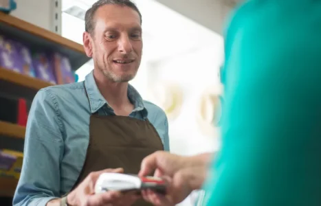 A shop owner in an apron smiling as a customer pays by card at the counter, the moment to ask for a Google review.